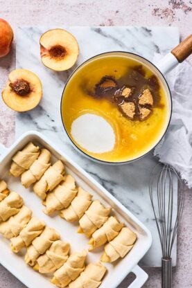 Crescent wrapped peach slices in a baking dish with a butter sauce being made in a pot on the side