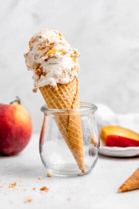 Two scoops of peach ice cream on a cone in a glass with peaches in the background