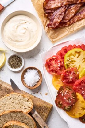 Slices of heirloom tomatoes on a white platter with aioli and bacon slices ready to build a sandwich