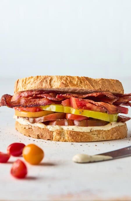 A bacon and tomato sandwich with garlic aioli on sourdough bread with small tomatoes in white background