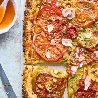 A summer tomato tart recipe with a slice cut ready to serve against a gray background and hot honey in a small bowl nearby