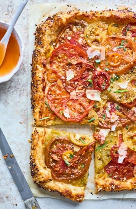 A summer tomato tart recipe with a slice cut ready to serve against a gray background and hot honey in a small bowl nearby