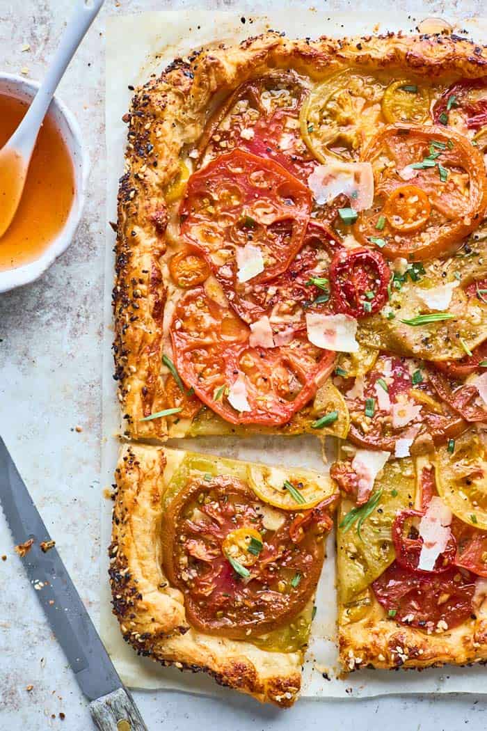 A summer tomato tart recipe with a slice cut ready to serve against a gray background and hot honey in a small bowl nearby