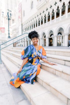 Jocelyn Delk Adams sitting on stairs in front of The Venetian in blue, black and orange dress