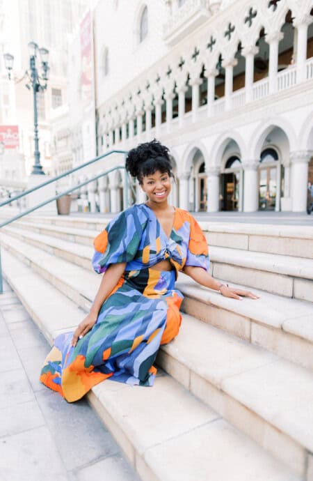 Jocelyn Delk Adams sitting on stairs in front of The Venetian in blue, black and orange dress