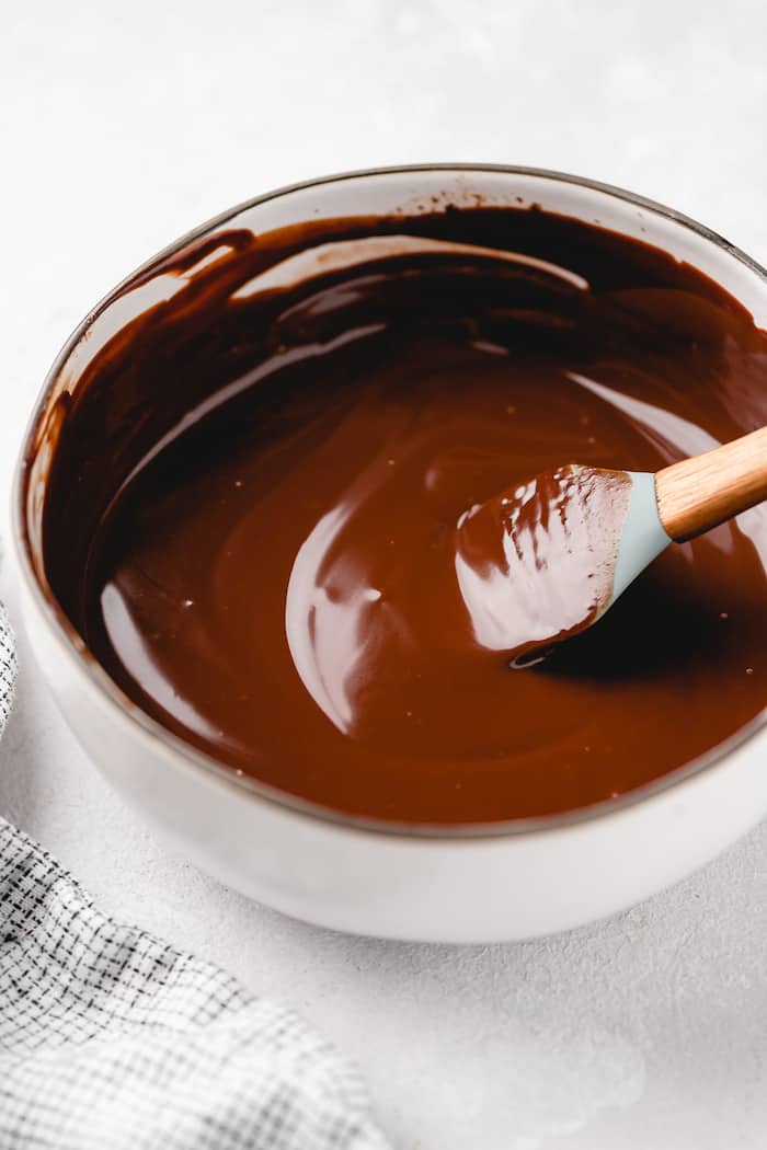 A spatula stirring a smooth chocolate ganache in a white pot on a white background