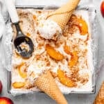 Homemade peach cobbler ice cream in a loaf pan with waffle cones, an ice cream scoop, sliced peaches, and whole peaches scattered around