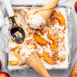 Homemade peach cobbler ice cream in a loaf pan with waffle cones, an ice cream scoop, sliced peaches, and whole peaches scattered around