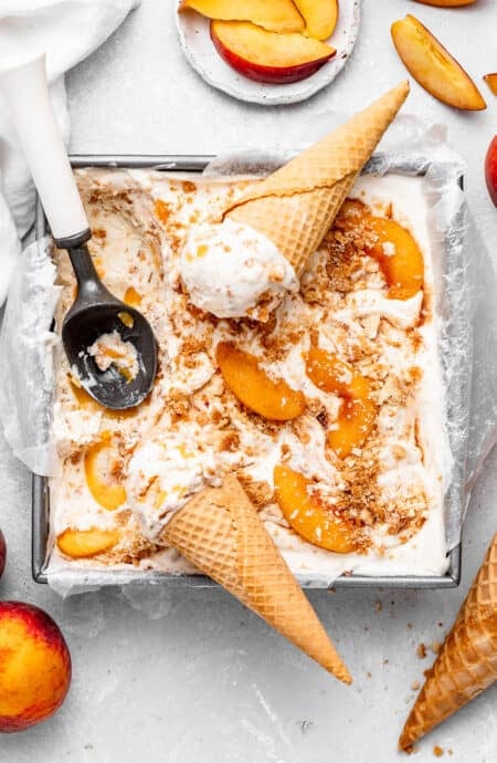 Homemade peach cobbler ice cream in a loaf pan with waffle cones, an ice cream scoop, sliced peaches, and whole peaches scattered around