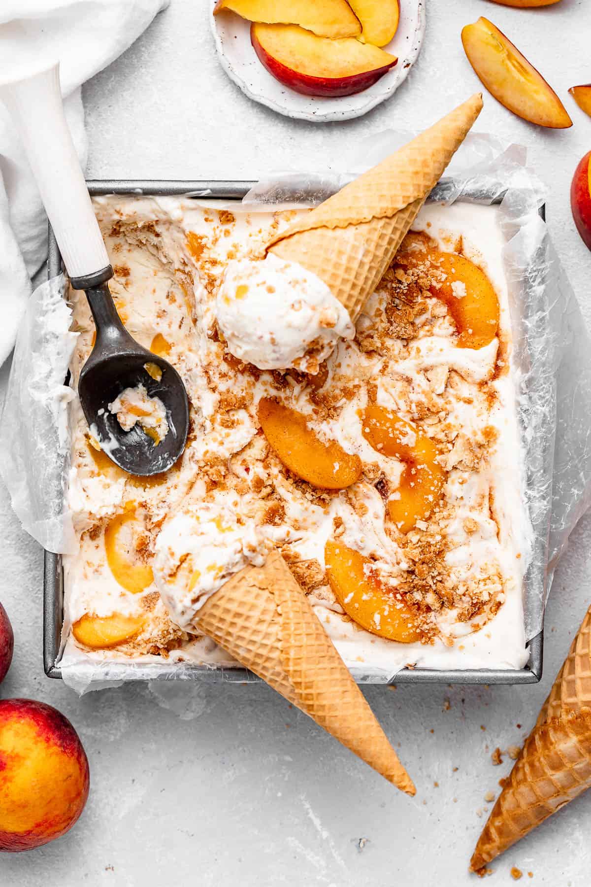 Homemade peach cobbler ice cream in a loaf pan with waffle cones, an ice cream scoop, sliced peaches, and whole peaches scattered around