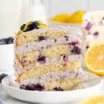 A single slice of blueberry lemon cake with visible blueberries and creamy frosting on a white plate, garnished with blueberries and lemon zest