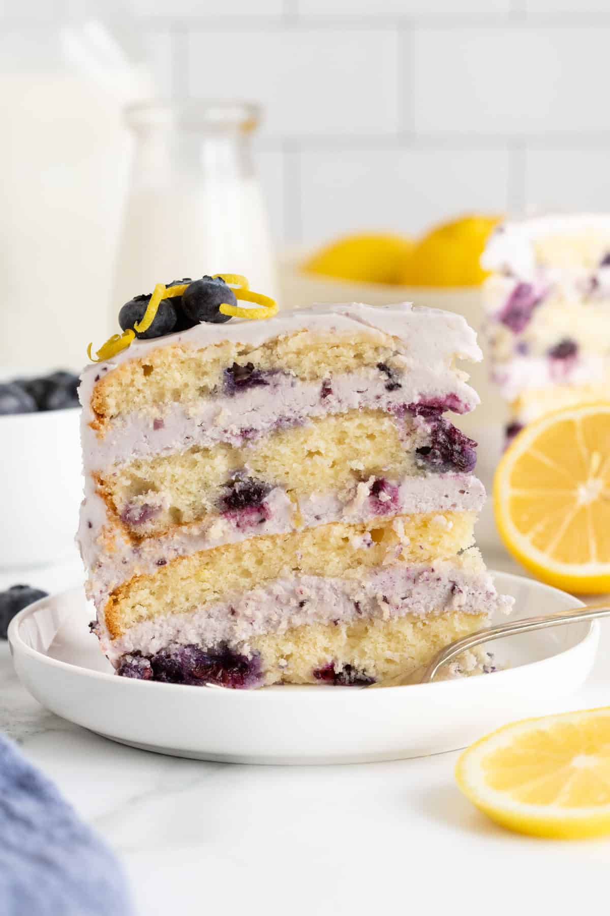 A single slice of blueberry lemon cake with visible blueberries and creamy frosting on a white plate, garnished with blueberries and lemon zest