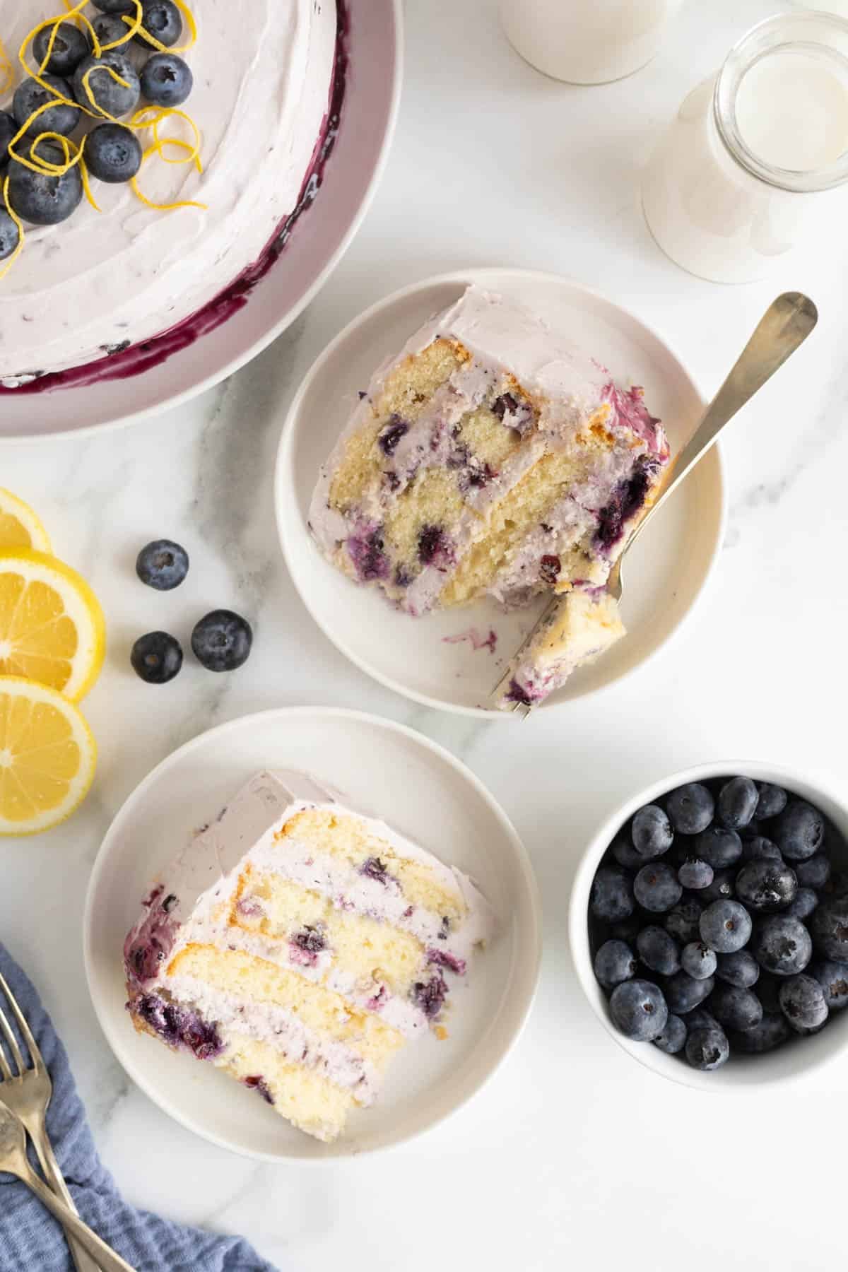 Overhead shot of two slices of blueberry lemon cake on white plates with a bowl of blueberries, milk bottles and lemon slices nearby