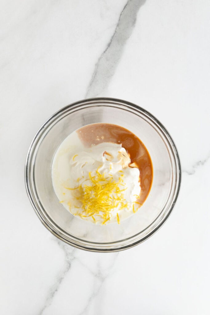 A glass mixing bowl with sour cream, lemon zest, and other wet ingredients