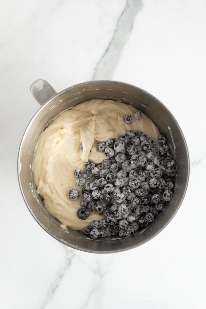 A metal mixing bowl with cake batter and a pile of fresh blueberries dusted with flour