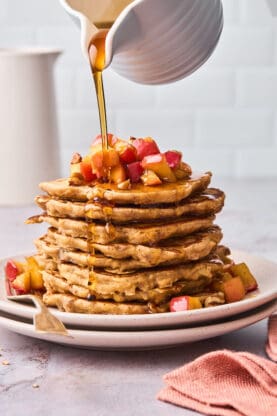 Maple syrup drizzling over a large stack of apple cinnamon pancakes ready to serve