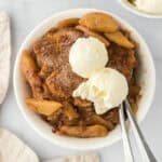 Two white bowls of apple cobbler recipe with scoops of vanilla ice cream on top with silver spoon on white background