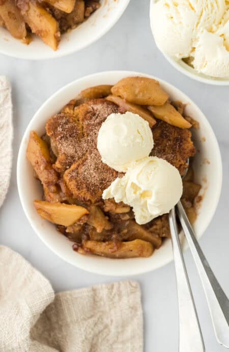 Two white bowls of apple cobbler recipe with scoops of vanilla ice cream on top with silver spoon on white background