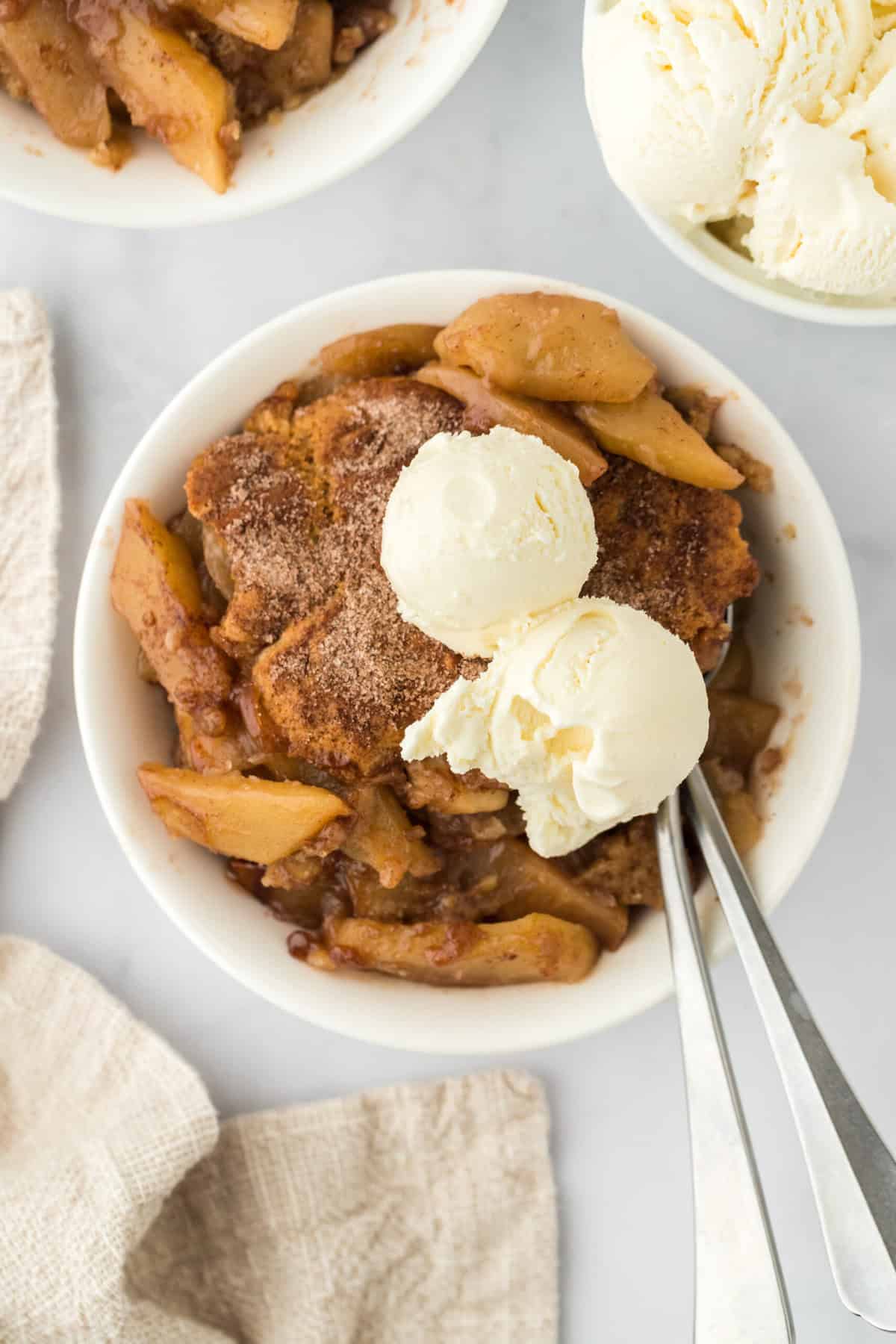 Two white bowls of apple cobbler recipe with scoops of vanilla ice cream on top with silver spoon on white background