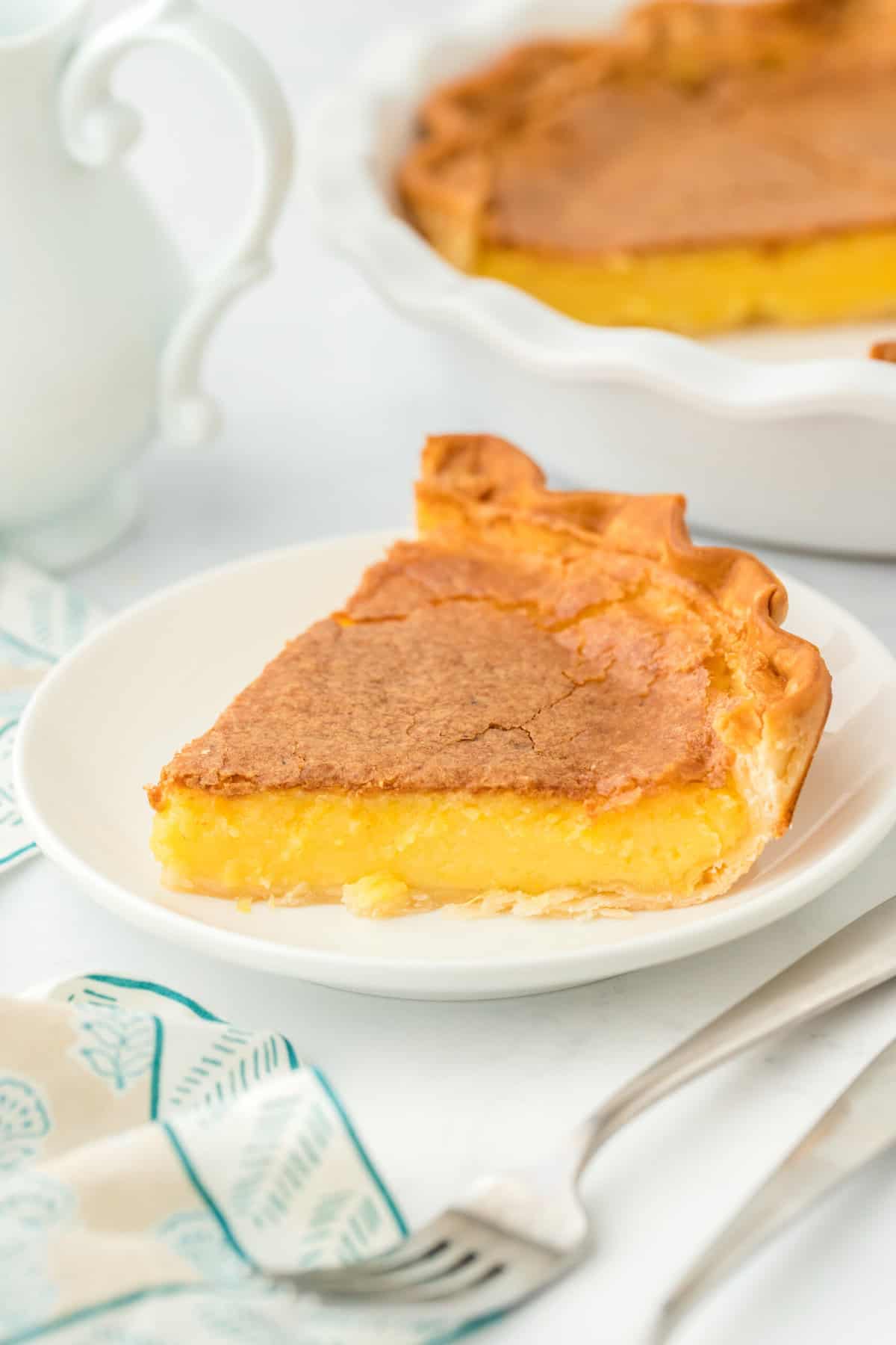 A slice of the best chess pie recipe on a white plate with striped napkin and full pie in white background