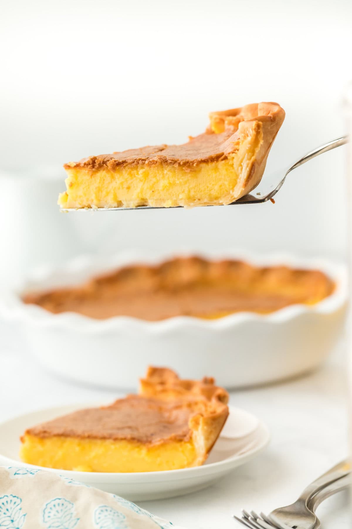 A slice of southern chess pie on a server lifted in the air with full pie in the white background