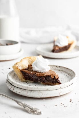 A slice of chocolate chess pie on a white speckled plate with whipped cream on top