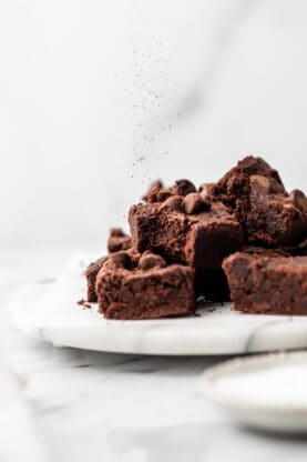 A stack of vegan brownies on a white dish with chocolate powder raining down