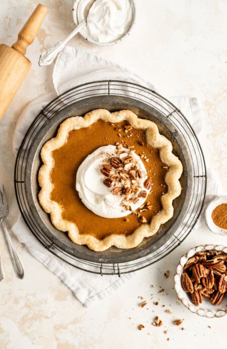 A classic pumpkin pie baked with whipped cream and pecans against a white background with a rolling pin next to it