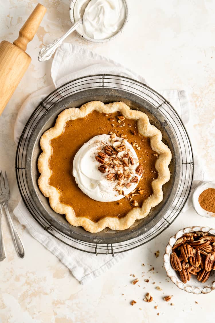 A classic pumpkin pie baked with whipped cream and pecans against a white background with a rolling pin next to it