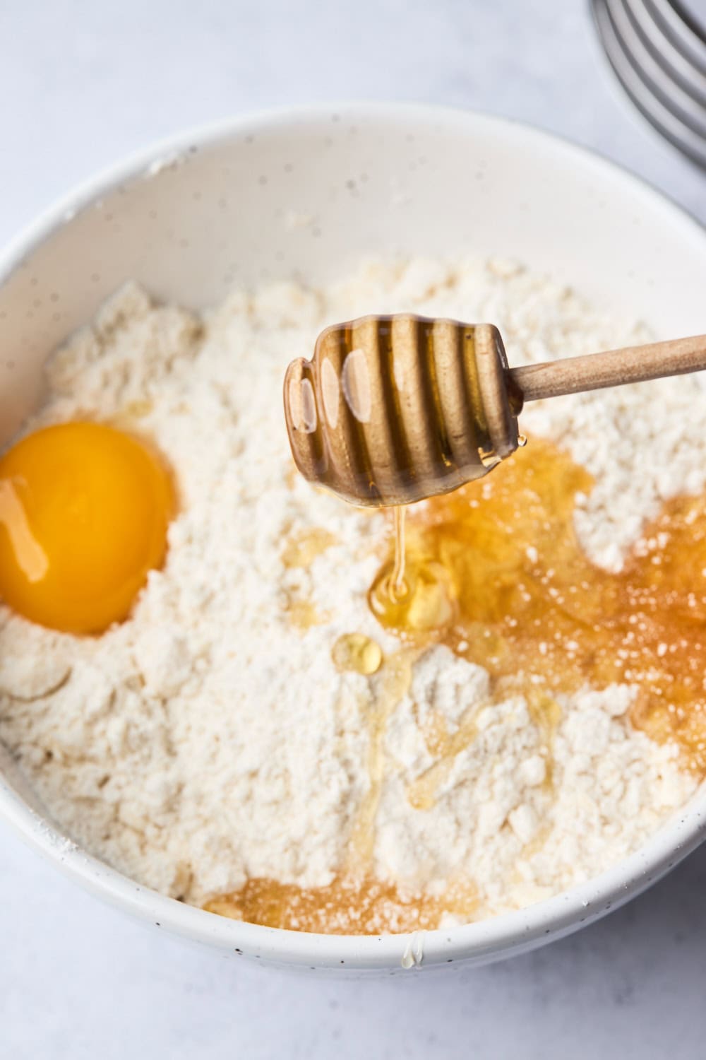 Close-up of a mixing bowl with flour, a cracked egg yolk, and honey being drizzled in with a honey dipper