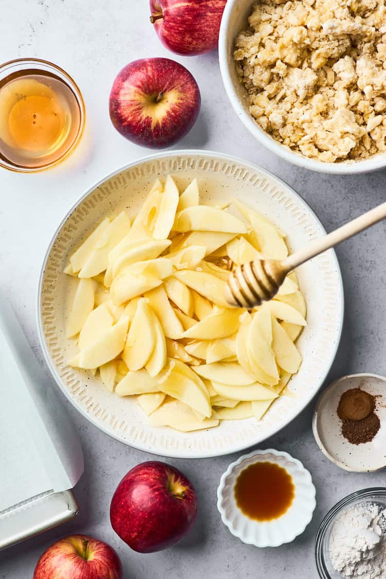 Bowl of peeled and sliced apples being drizzled with honey, surrounded by baking ingredients like cinnamon, vanilla, and whole red apples