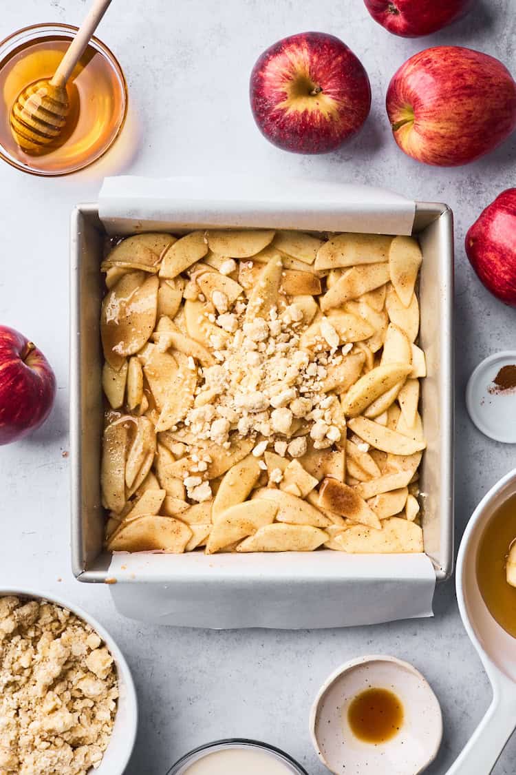 Unbaked apple bars in a parchment-lined square pan topped with sliced apples and crumble, with apples and ingredients scattered around