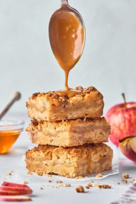 Honey caramel sauce dripping down over a stack of apple crumble bars against a gray background