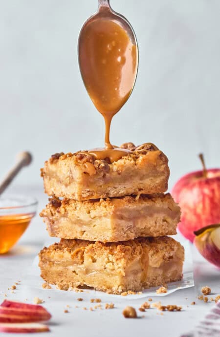 Honey caramel sauce dripping down over a stack of apple crumble bars against a gray background