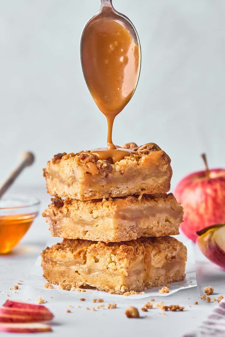 Honey caramel sauce dripping down over a stack of apple crumble bars against a gray background