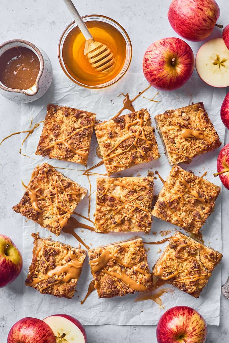 An overhead of 9 apple bars with caramel sauce drizzled on top and a bowl of honey with a drizzler nearby and apples in the background