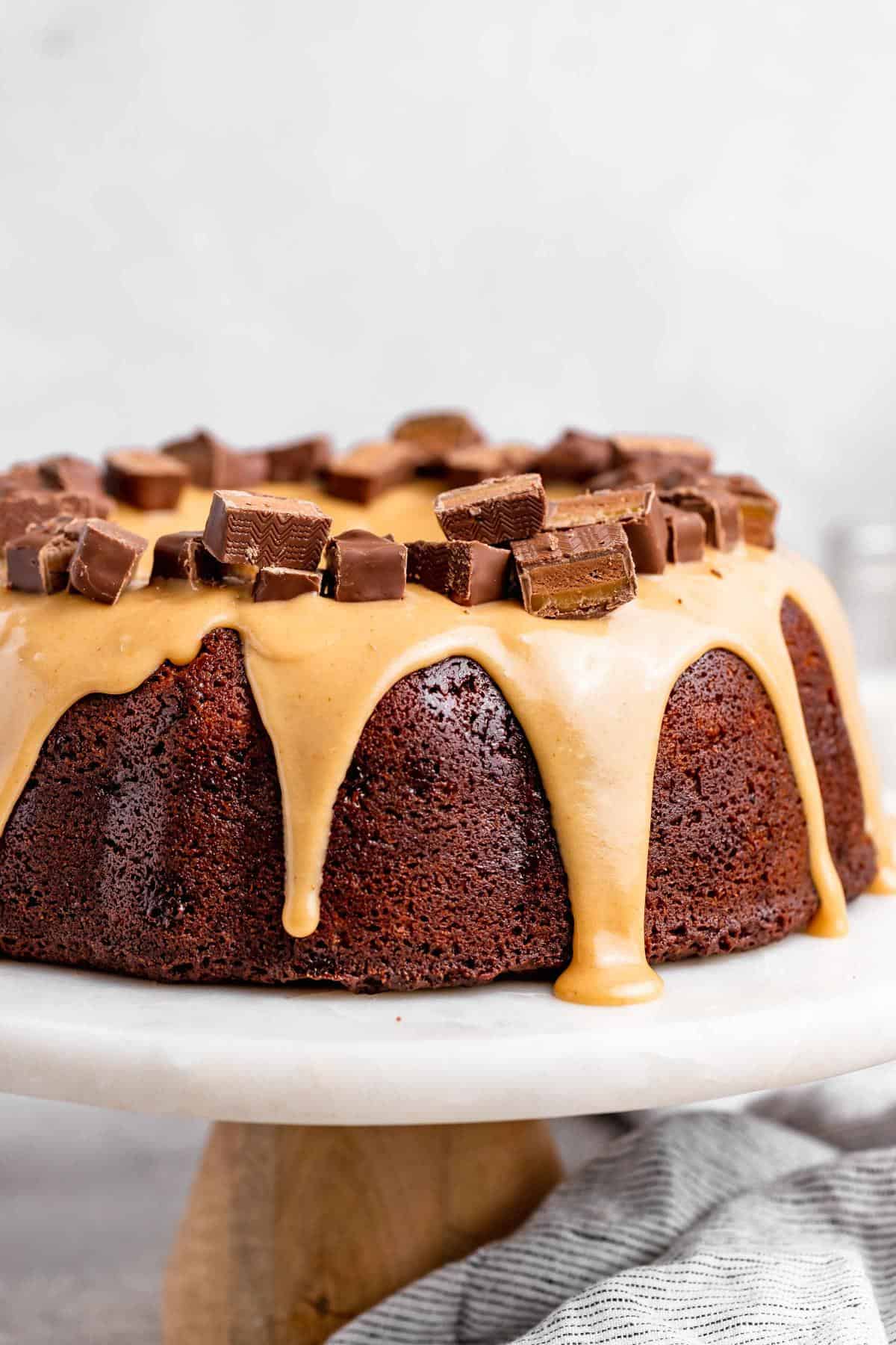 A milky way bundt cake drizzled with caramel glaze and topped with chopped milky way candy bars on a white cake stand