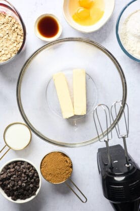 Butter in a clear bowl with ingredients for cookies nearby and a hand mixer