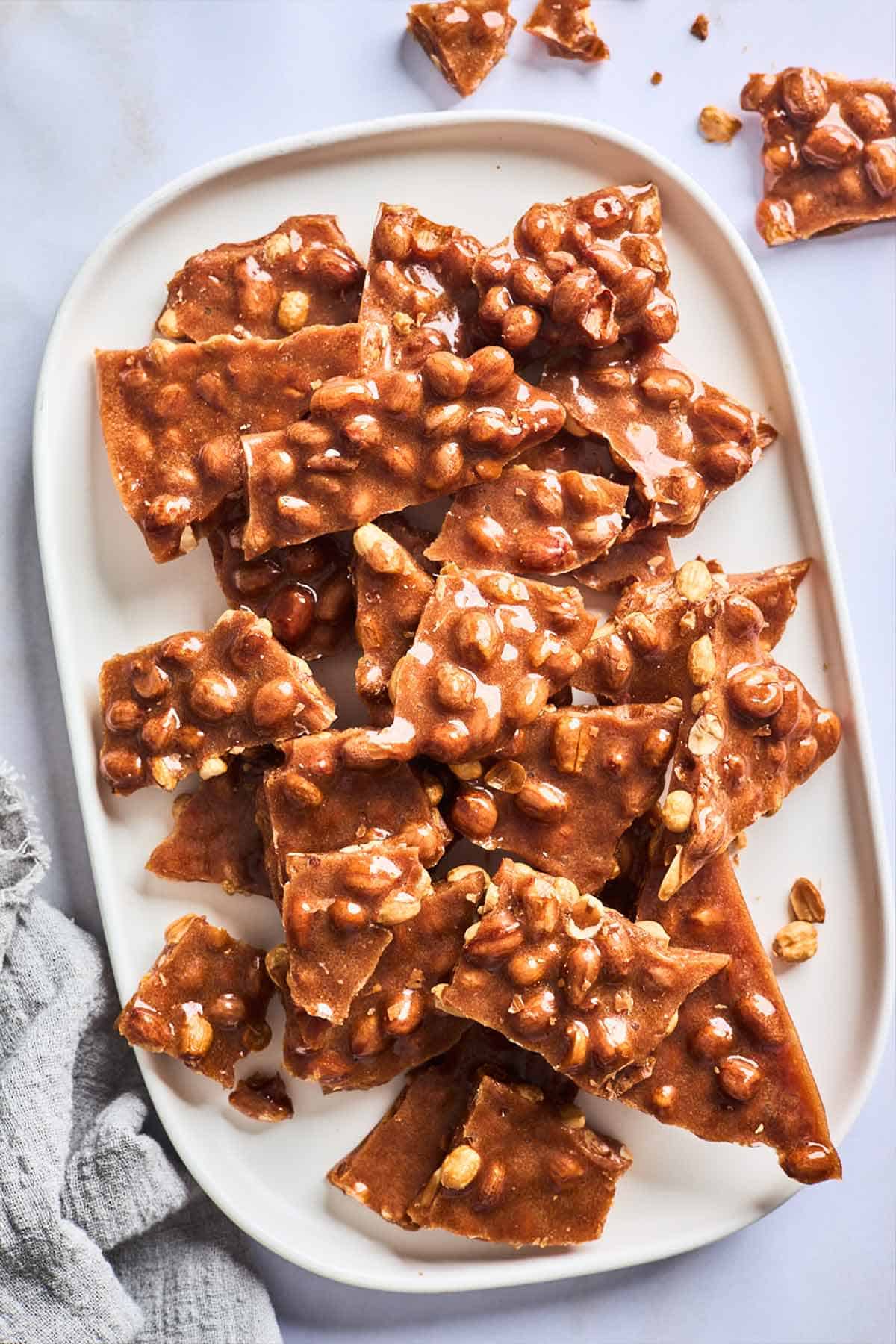 An oval white serving tray loaded up with peanut brittle with a few pieces scattered on the table next to it.