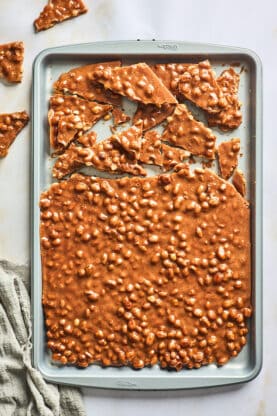 Some peanut brittle being cracked and broken apart on a large baking sheet