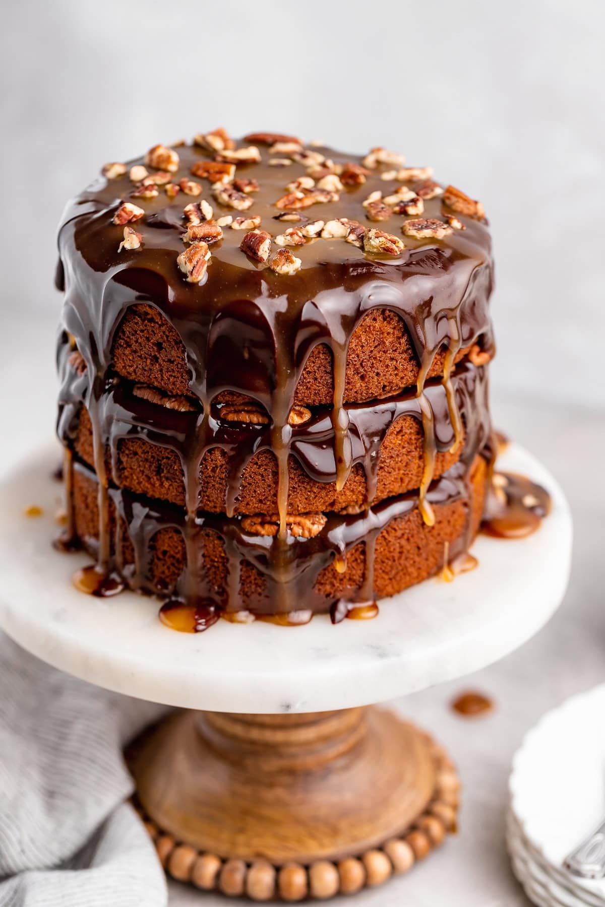 Full view of a three layer pumpkin spice turtle cake topped with chocolate, caramel, and pecans, sitting on a wooden pedestal cake stand