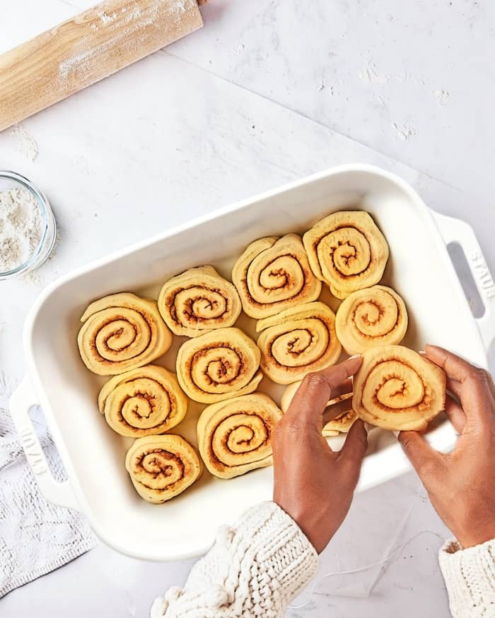 Two hands placing cinnamon rolls in white baking dish