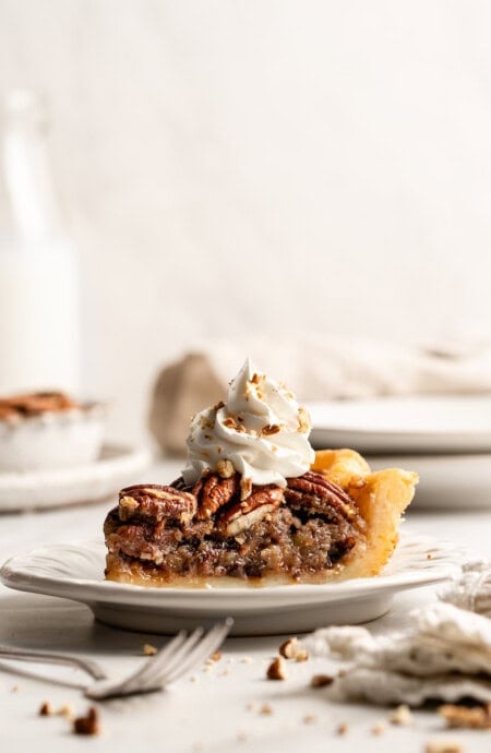 A slice of chocolate pecan pie sitting on a white plate with whipped cream on top against white background