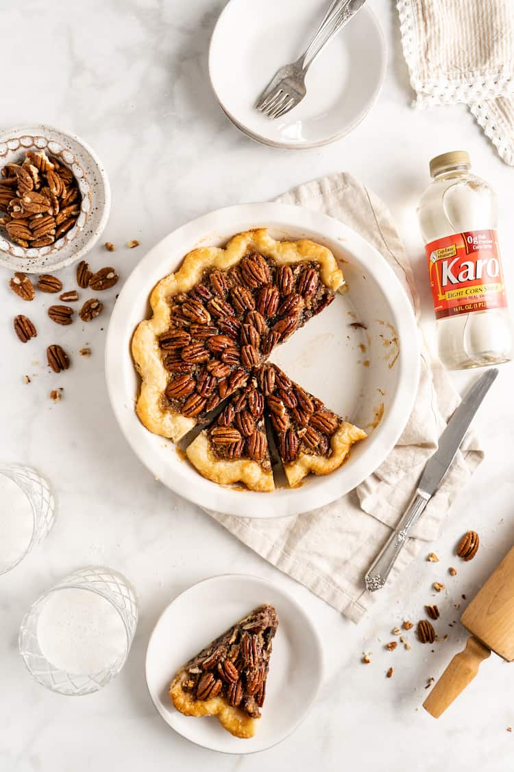 An overhead shot of a sliced Chocolate Bourbon Pecan Pie with pecans in white background and a slice on a plate and corn syrup in bottle
