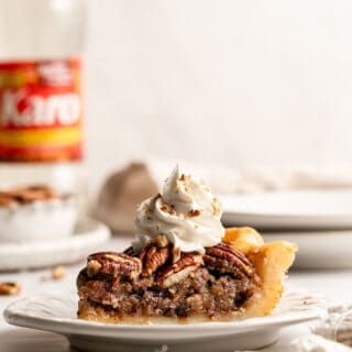 A slice of chocolate pecan pie on a white plate against a white background with whipped cream and corn syrup in background