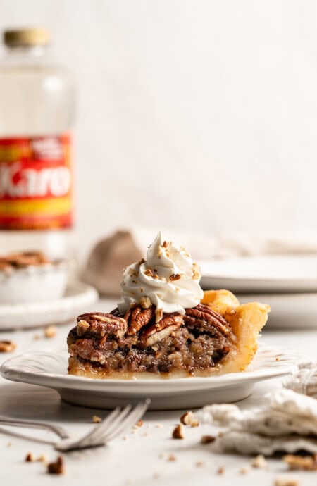 A slice of chocolate pecan pie on a white plate against a white background with whipped cream and corn syrup in background
