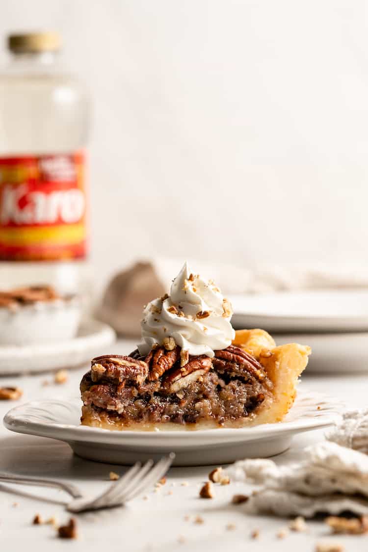 A slice of chocolate pecan pie on a white plate against a white background with whipped cream and corn syrup in background
