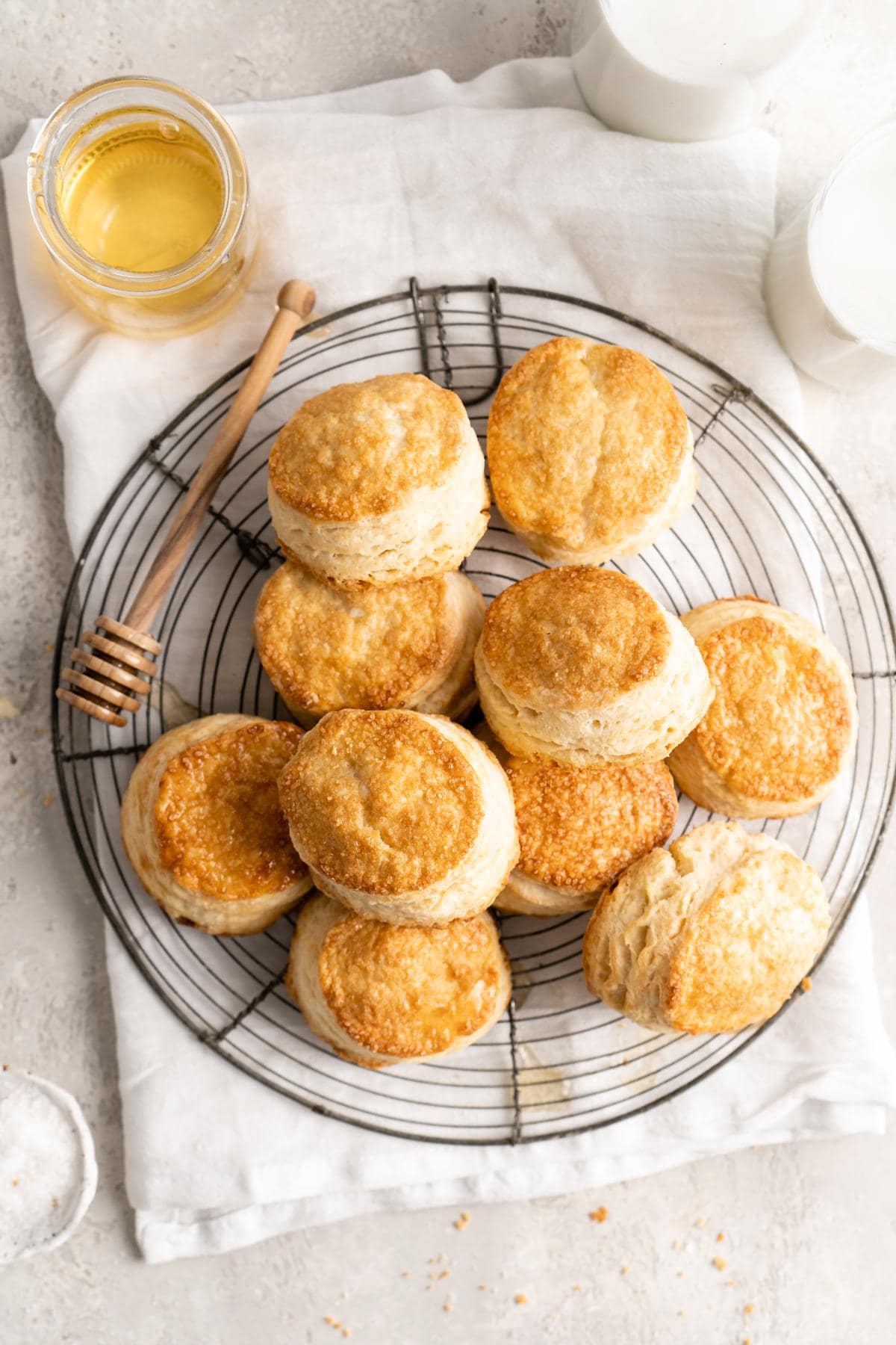 Overhead shot of a dozen golden biscuits arranged on a cooling rack with a honey jar and glasses of milk
