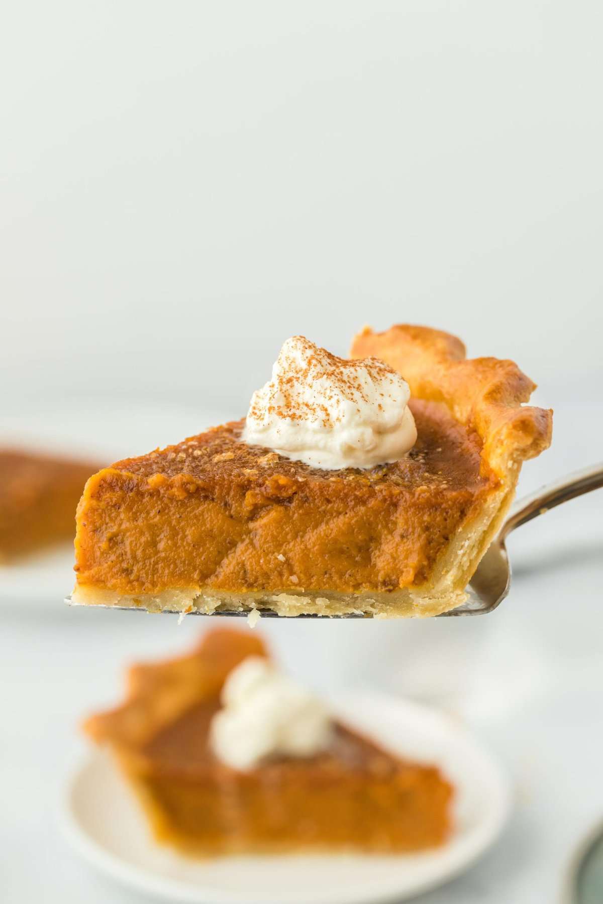 A pie server holding a slice of pumpkin pie with whipped cream on top, with another slice of pie on the background