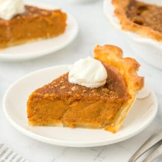 A slice of pumpkin pie on a plate topped with whipped cream with a fork next to it and more slices on the background